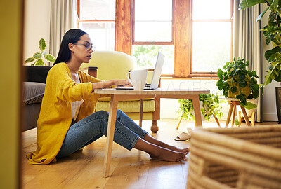 Focused woman working remotely sitting on the floor at a coffee table in a living room. A serious young hispanic female wearing glasses typing on her laptop at home. Using technology during lockdown