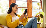 Hispanic woman using her smartphone sitting in front of a window in a bright living room. A young happy female smiling at her cellphone relaxing on a chair using modern technology at home in lockdown