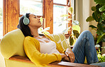 Woman listening to music on headphones while comfortable and relaxing in a bright living room. A young hispanic female with eyes closed resting and sitting on a chair using modern technology at home