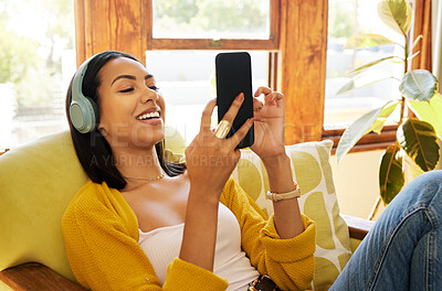 Hispanic woman using her smartphone and listening to music on headphones while comfortable and relaxing in a bright living room. A young female sitting on a chair texting on her cellphone at home