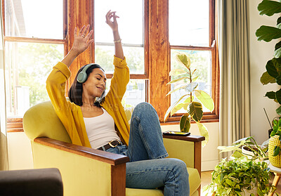 Hispanic woman listening to music on headphones while dancing and vibing in a bright living room. A young female with eyes closed relaxing and sitting on a chair using modern technology at home