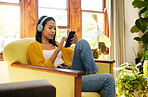 Woman using her smartphone and listening to music on headphones while comfortable and relaxing in a bright living room. A young female sitting on a chair texting on her cellphone at home