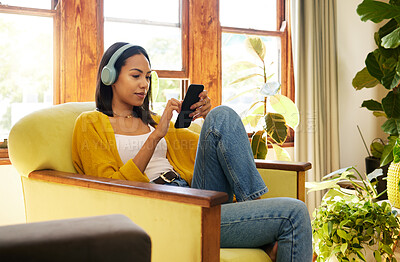Woman using her smartphone and listening to music on headphones while comfortable and relaxing in a bright living room. A young female sitting on a chair texting on her cellphone at home