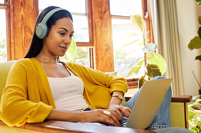Hispanic woman typing on a laptop and listening to music on headphones, smiling and working in a bright living room. A young female sitting on a chair using modern technology at home in lockdown