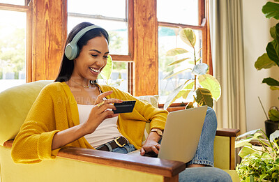 Woman shopping online while typing on her laptop and holding her bank card sitting on a chair in a bright living room in front of a window. A young female at home using modern technology