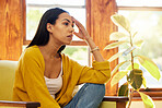 Stressed woman at home sitting on a chair in a bright living room with her hand against her head infront of a large window. One worried and anxious young female inside with headache over debt