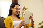 Hispanic woman using her tablet while relaxing in a bright living room. A young female with glasses sitting on a sofa scrolling on her digital tablet at home using modern technology during lockdown