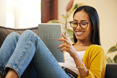 Woman using her tablet while comfortable and smiling in a bright living room. A young female with glasses relaxing on a sofa looking at her digital tablet at home using modern technology