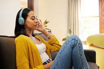 Hispanic woman listening to music on headphones while comfortable and relaxing in a bright living room. A young female with eyes closed resting and sitting on a sofa using modern technology at home