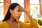Worried woman at home sitting on a chair in a bright living room with her hand against her head holding a smartphone. One tired and anxious young female inside with a headache and stress