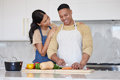 A young hispanic couple wearing aprons cooking together at home. Latin man cutting carrots and other vegetables while his girlfriend holds him and smiles