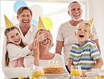 Little girl covering mother's eyes while celebrating her birthday and surprising her. Happy woman having party at home with her two children, husband and father, wearing party hats and laughing