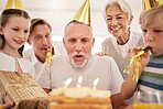 Senior man celebrating his birthday with his family at home, wearing party hats and blowing whistles. Grandpa blowing out birthday candles and making a wish surrounded by his grandkids, wife and son