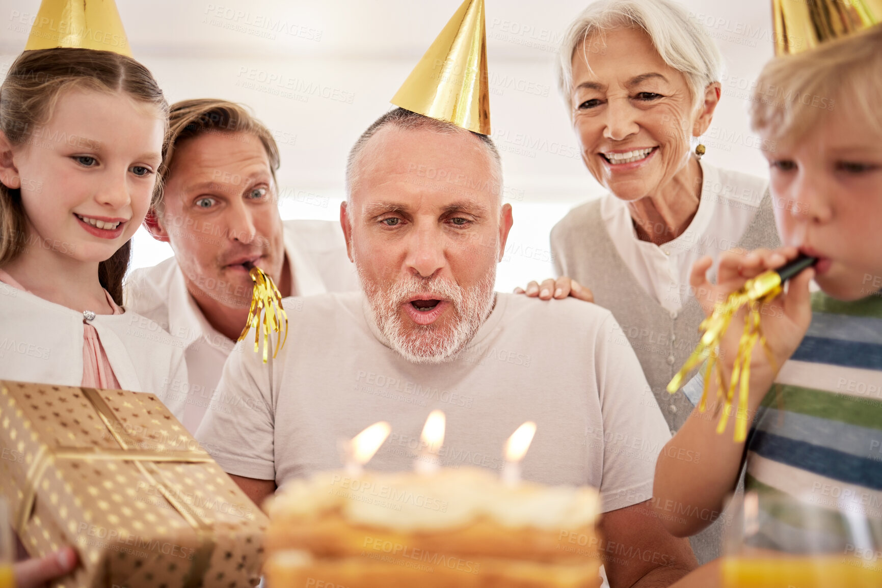 Buy stock photo Senior man celebrating his birthday with his family at home, wearing party hats and blowing whistles. Grandpa blowing out birthday candles and making a wish surrounded by his grandkids, wife and son