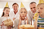 Senior woman celebrating her birthday with family at home, wearing party hats and blowing whistles. Grandma looking at birthday cake and being joyful while surrounded by her grandkids, husband and son