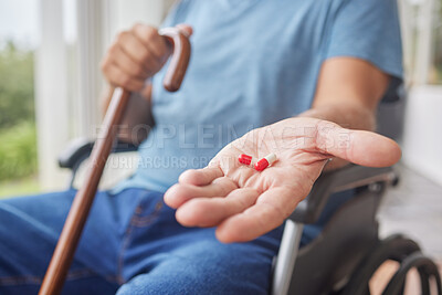 An unknown man in a wheelchair showing his pills while holding a walking stick and sitting at home. Unrecognizable mature man taking his daily treatment for chronic disease and illness