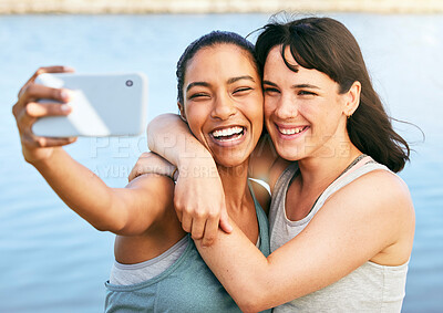 Two female friends taking selfies after a workout in nature and standing in front of a lake using a smartphone. Young mixed race female using her phone to take a selfie of herself and her caucasian friend