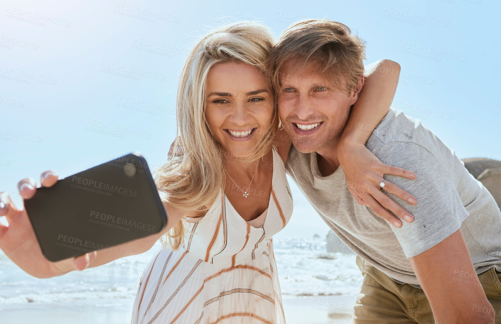 Buy stock photo Loving young couple taking selfie with phone while enjoying a day at the beach smiling and hugging. Romantic husband and wife showing affection and taking picture for social media while on vacation