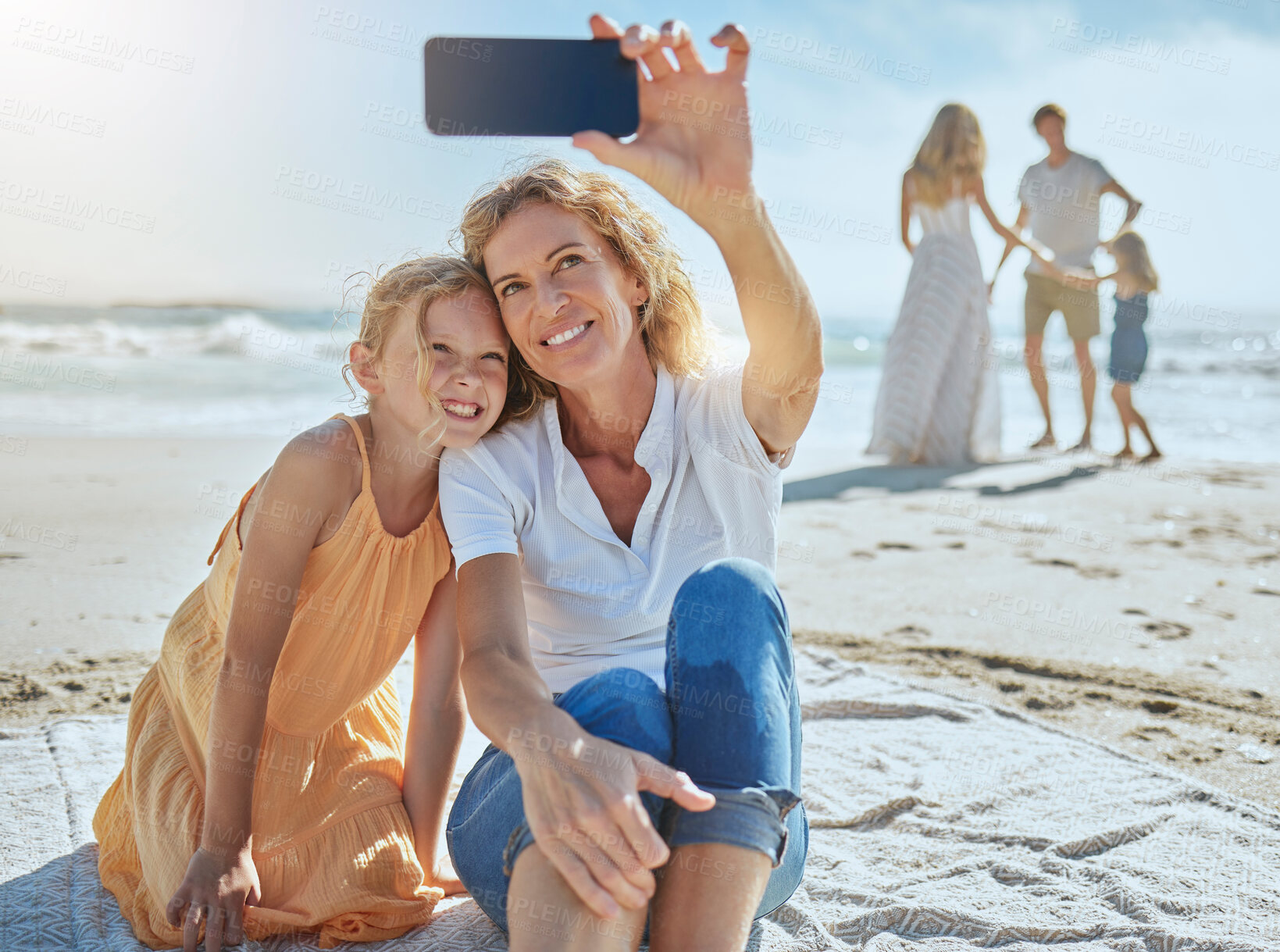 Buy stock photo Cheerful mature woman and little girl taking a selfie while sitting on the beach. Happy little girl smiling while sitting with her mom or grandmother taking picture on mobile phone while on vacation
