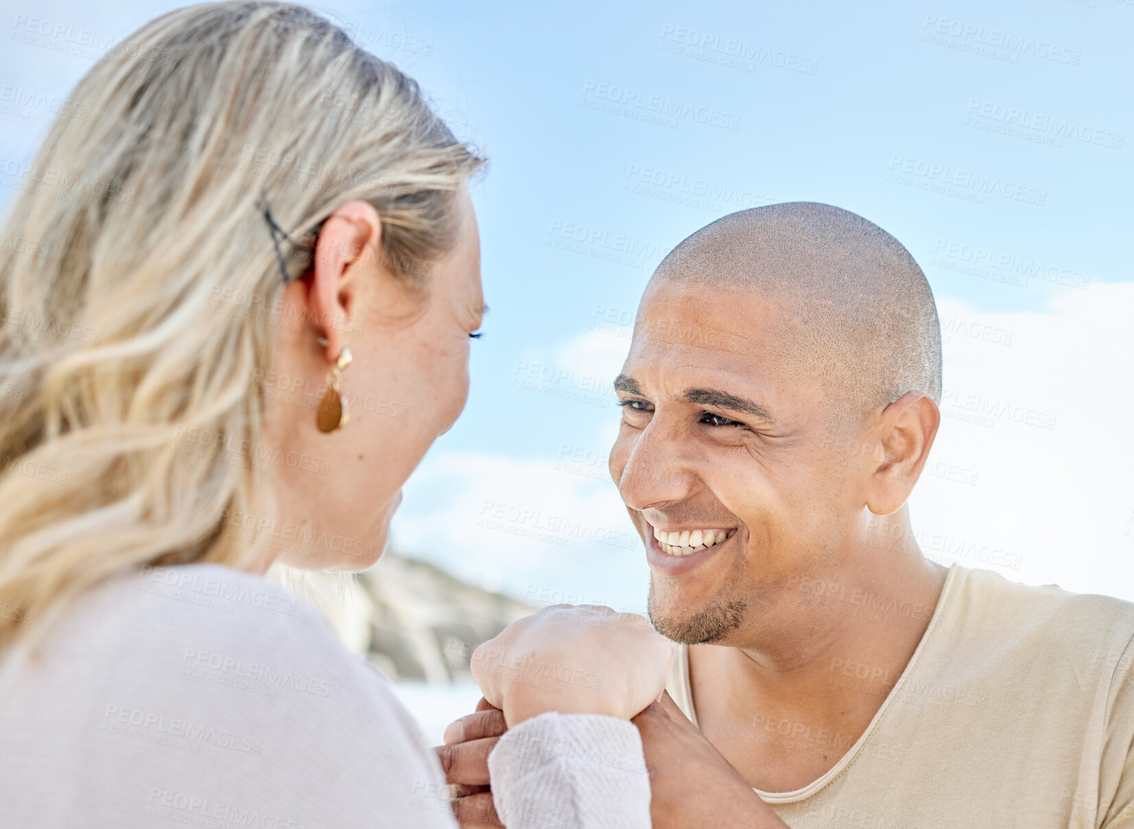 Buy stock photo A young man proposing to his girlfriend at the beach in summer. Happy interracial couple holding hands and smiling. Soon to be husband and wife staring lovingly into one another's eyes outside