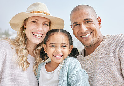 Buy stock photo Portrait of an interracial happy family bonding while on a beach vacation together. Cute little girl embracing and sitting with her parents outside, enjoying fresh summer air with copyspace 