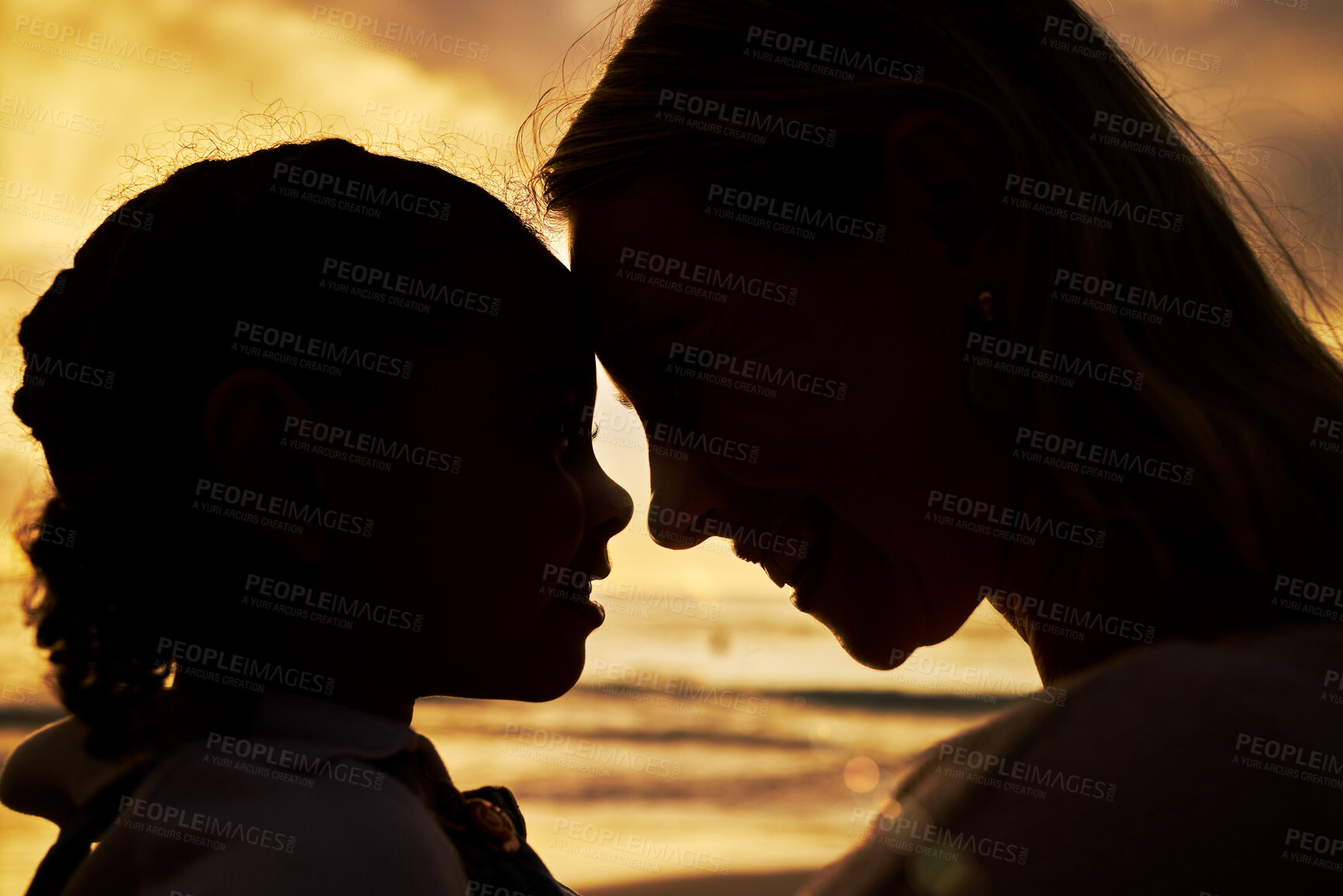 Buy stock photo Closeup silhouette of a mother and daughter standing face to face on the beach at sunset. Backlit young woman and her girl child smiling and facing one another with the ocean in the background