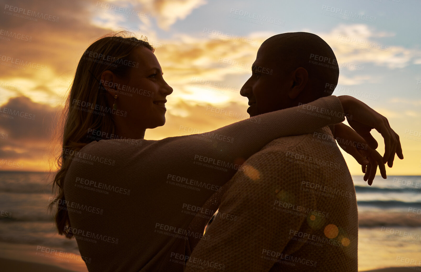 Buy stock photo Loving young affectionate interracial couple in silhouette facing each other while spending time together on the beach at sunset. Lovers sharing romantic moment against golden sky while on vacation