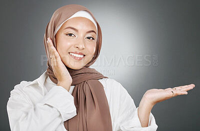 Buy stock photo Portrait of glowing beautiful muslim woman isolated against grey studio background. Young woman wearing a hijab or headscarf showing flawless skin and holding her hand out for a product or treatment 