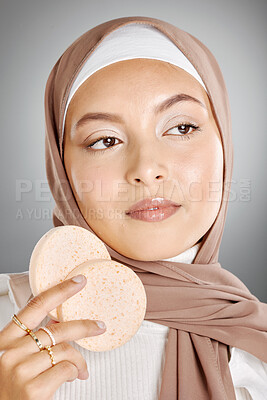 Buy stock photo Beauty portrait of an attractive serious sensual young Muslim woman standing wearing a beige hijab and holding two sponge beauty blenders against gray studio background. A girl with brown lipstick on her lips.