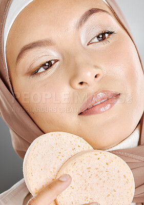 Buy stock photo Beautiful muslim woman holding and using makeup sponges on her face against a grey studio background. Young female wearing an hijab and doing her beauty skincare routine. Taking care of healthy skin
