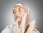 One beautiful young muslim woman wearing brown headscarf posing with pampas wheat plant against grey studio background. Modest female arab wearing makeup with face covered in traditional hijab