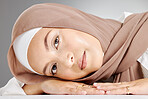 Studio portrait of one beautiful young muslim woman wearing brown headscarf lying against grey background. Happy arab female wearing makeup with face covered in hijab for traditional modesty