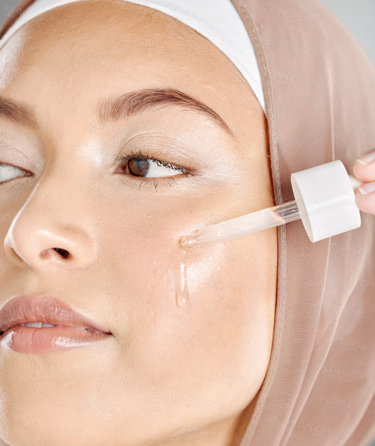 Buy stock photo A young woman applying antiaging facial serum to her face and skin. Beautiful muslim girl wearing a hijab trying retinol with a dropper as her skincare routine and beauty treatment regime