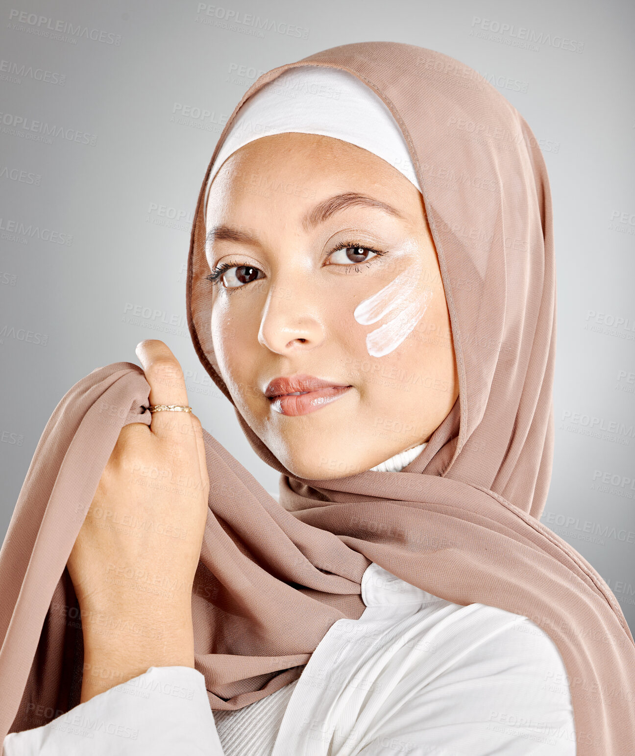 Buy stock photo Portrait of a Muslim woman using face moisturiser, sunscreen or lotion isolated on a grey studio background with copyspace. Young female wearing a hijab or headscarf while doing her skincare routine