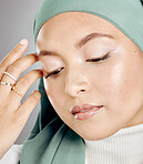 Closeup of a glowing beautiful muslim woman isolated against grey studio background. Young woman wearing a hijab or headscarf showing her eyelash extensions and jewellery. Touching flawless skin