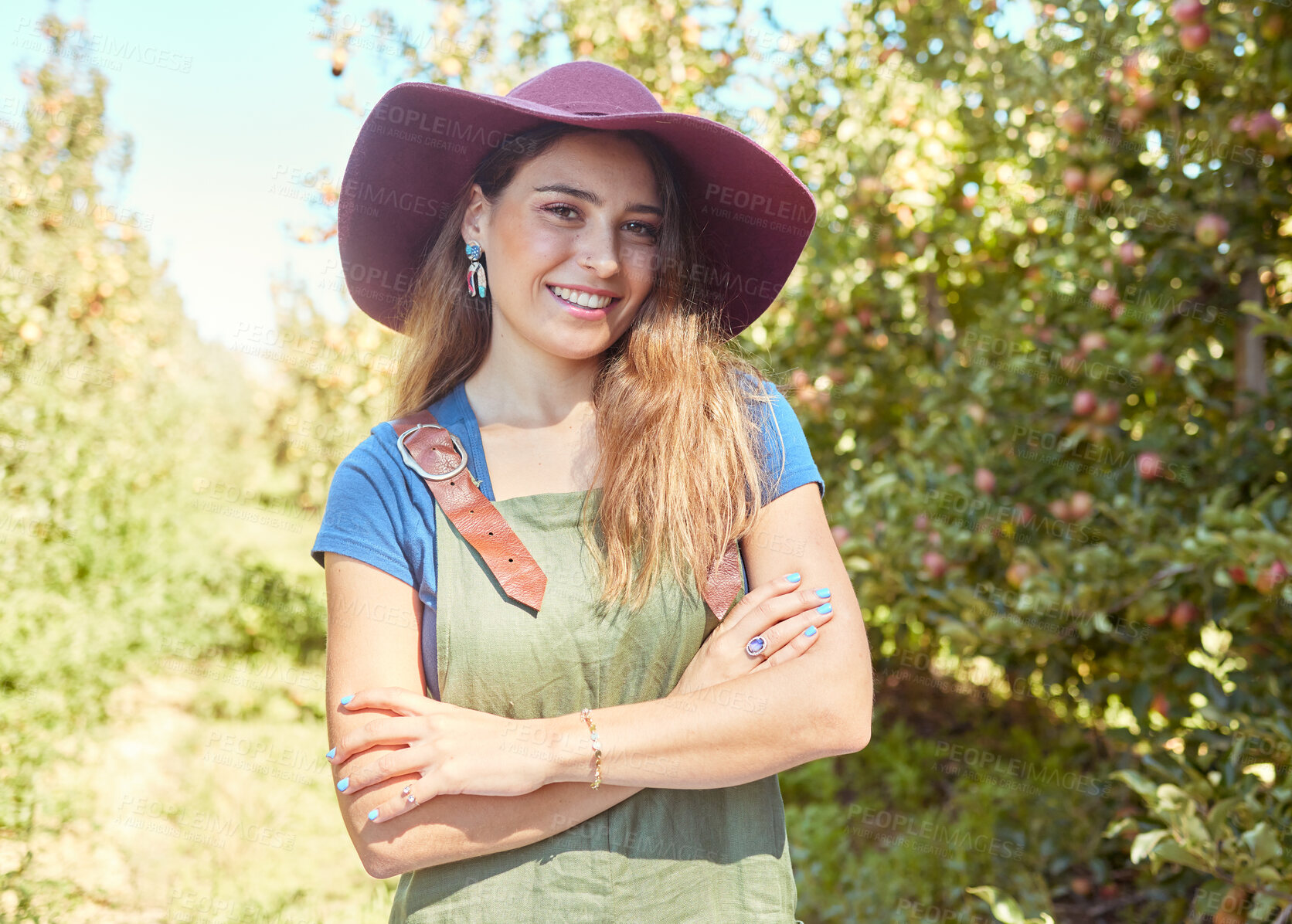 Buy stock photo A beautiful female farm worker standing with her arms crossed on a fruit farm during harvest season. Young happy farmer between fruit trees in summer. Agricultural industry growing produce