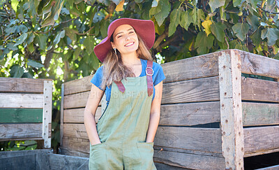 Portrait of a woman in a straw hat standing under a tree next to a rustic wooden crate. One young happy female wearing a summer hat and dungaree dress in a garden on a sunny day picking apples
