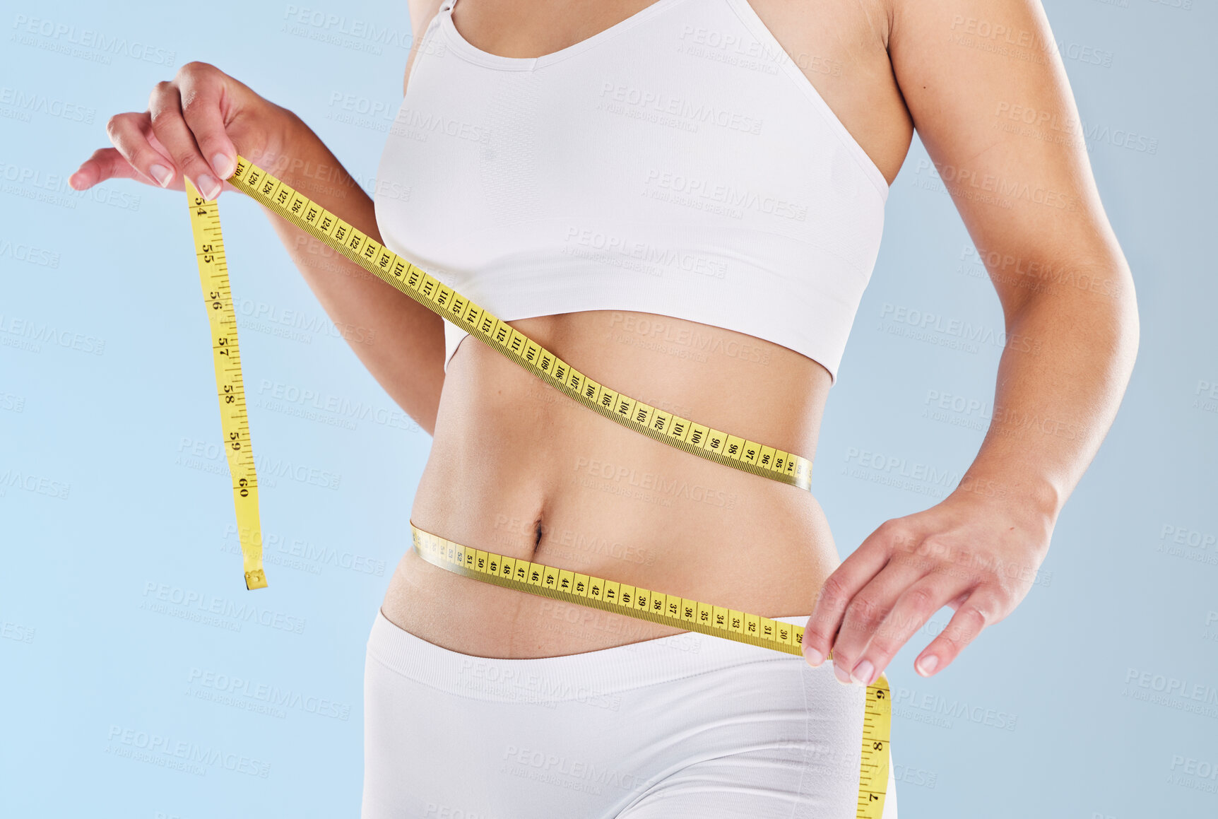 Buy stock photo A woman using a measuring tape to measure her flat stomach against a isolated blue studio background. Closeup hands of a female taking waist measurements to track weight loss progress while on a diet