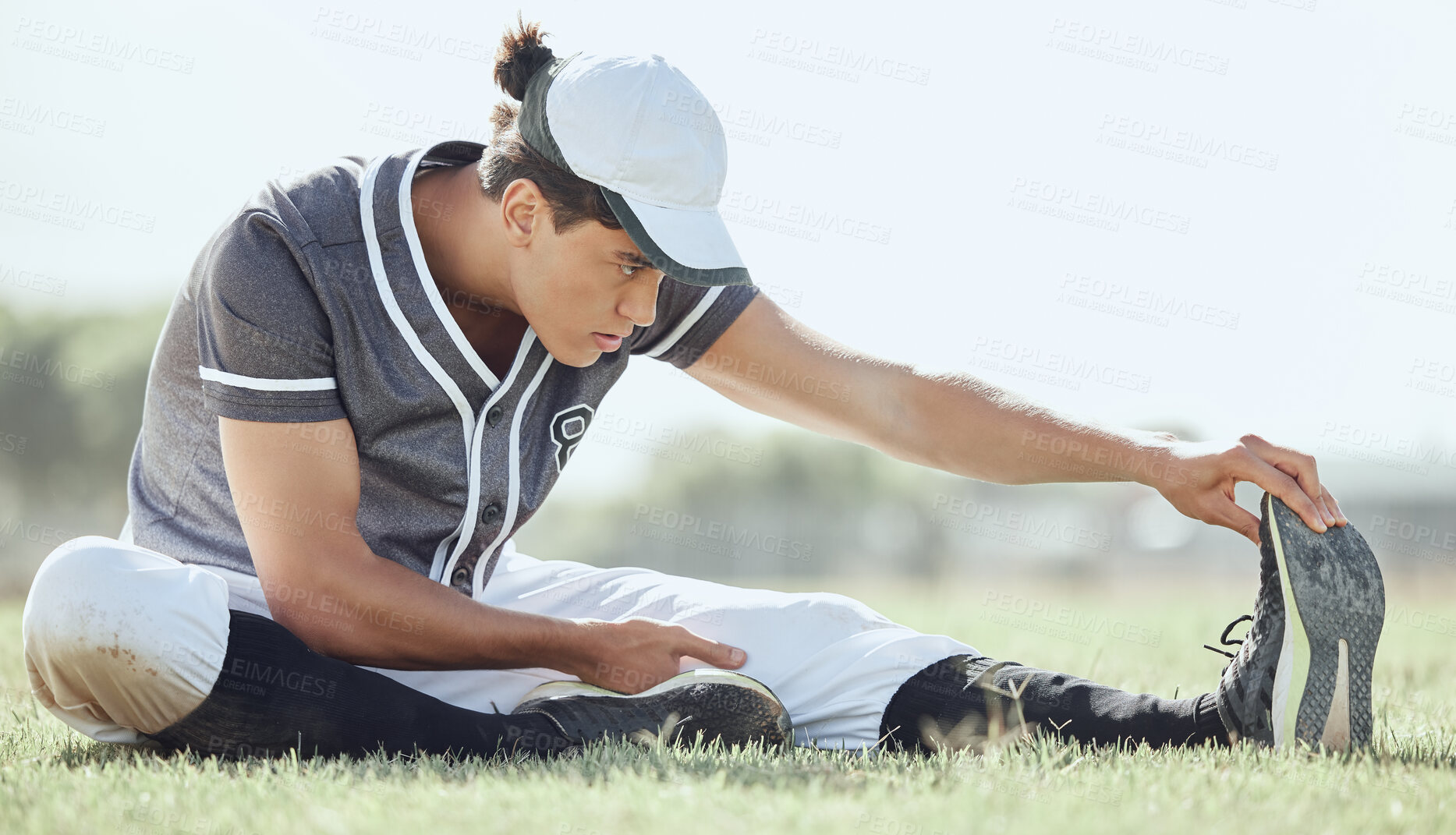 Buy stock photo Full length of a baseball player sitting on a pitch and stretching before playing a game. Serious and focused athlete getting ready to play match on grass. Fit, active, sporty, athletic man