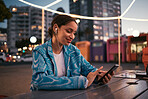 Happy, urban and trendy woman browsing on phone while wearing earphones and listening to music while sitting at outdoor cafe in night city. Happy woman streaming subscription app or making video call