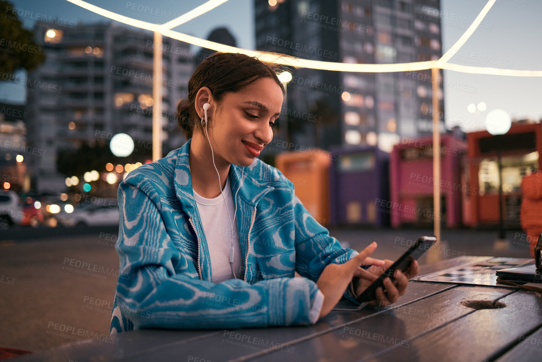Buy stock photo Happy and trendy woman browsing on phone while wearing earphones and listening to music while sitting at outdoor cafe in night city. Happy woman streaming subscription app or making video call