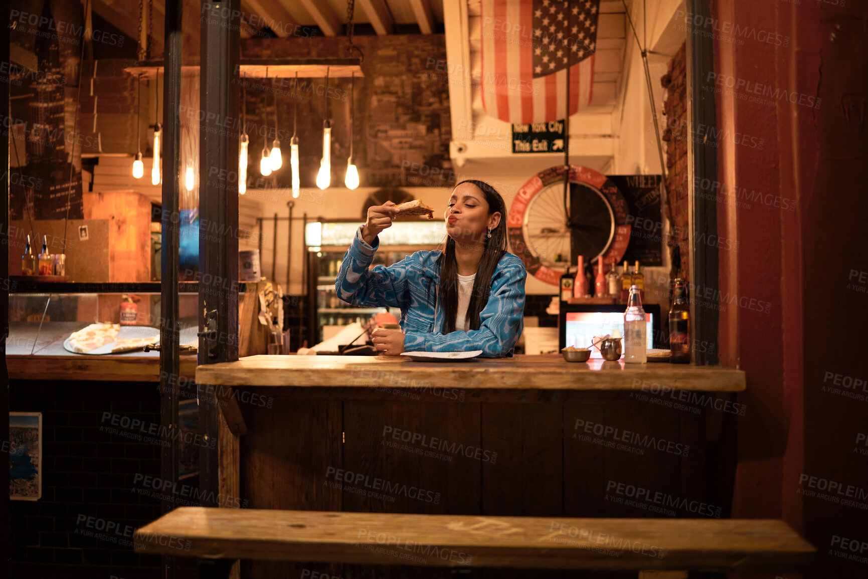 Buy stock photo A young and happy woman eating pizza sitting alone in an empty and dark American restaurant. A female relaxing and enjoying delicious food late at night in a cosy junk food, takeaway cafe