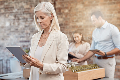 Buy stock photo Mature female architect using a tablet to analyze designs in a modern office. Senior business woman with a blurred background of a young architectural team working together on a building model.