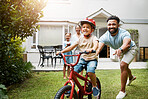 Learning, bicycle and proud dad teaching his young son to ride while wearing a helmet for safety in their family home backyard. Active father helping and supporting his child while cycling outside