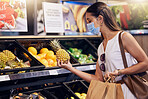 Shopping, checking and holding a fruit with a female checking if its good, ripe and healthy to eat. Young woman with a mask buying fresh and organic food at a retail store or supermarket indoors