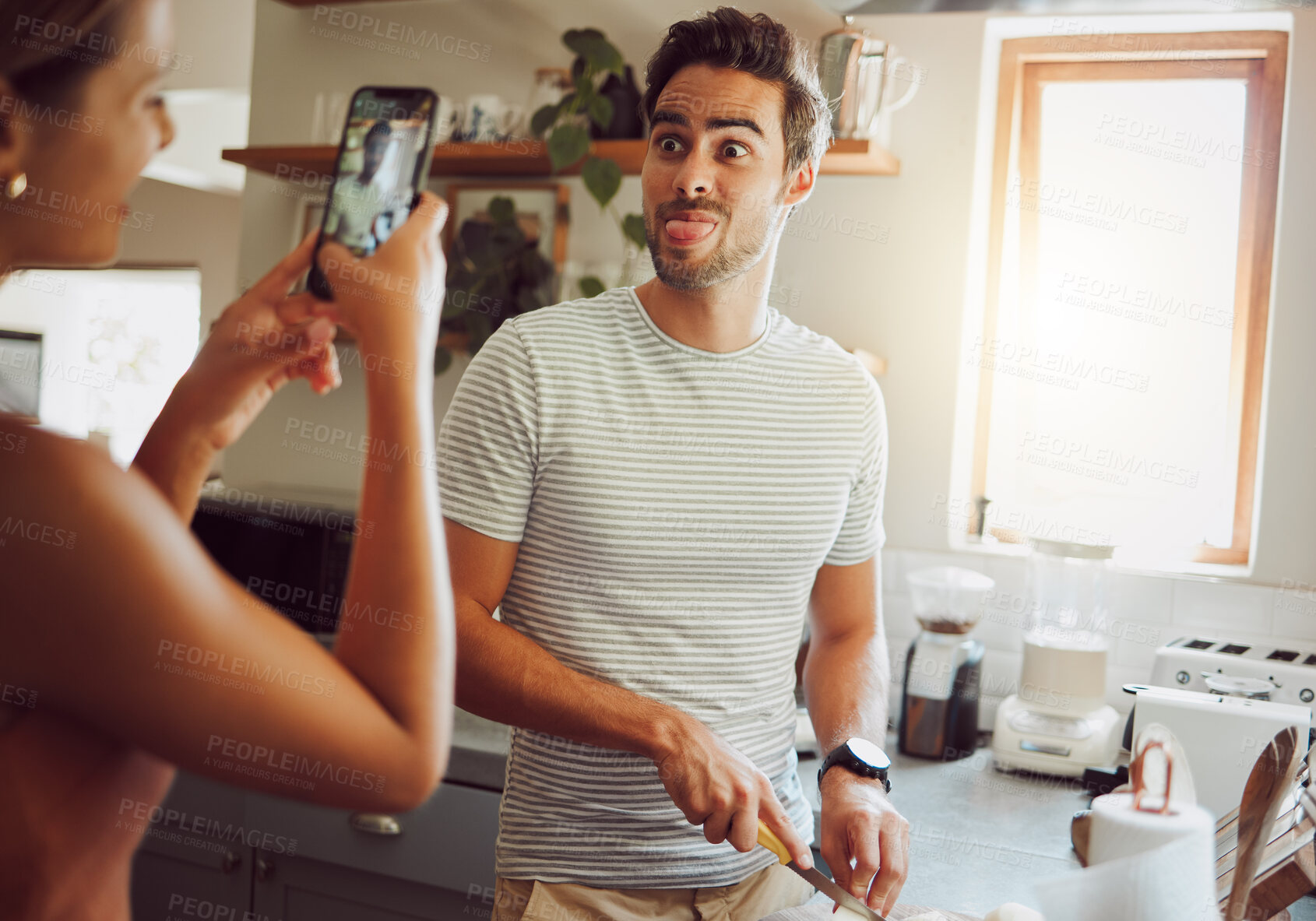Buy stock photo Silly, goofy and interracial couple taking pictures, having fun and cooking together in the kitchen at home. Happy girlfriend taking pictures of boyfriend, making funny faces and preparing dinner