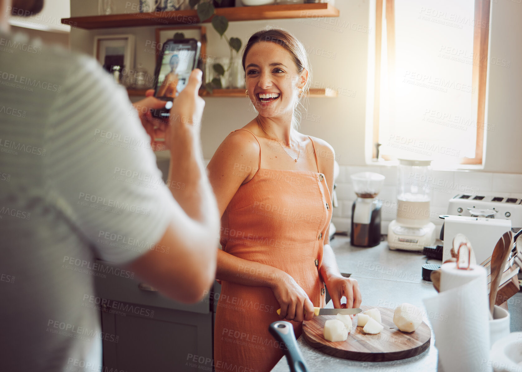 Buy stock photo Woman cooking with husband taking a picture in kitchen on his mobile phone at home. Beautiful, happy and female chef cutting vegetables to cook healthy, delicious and vegetarian food at a table. 