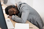 Tired CRM call center agent sleeping on computer desk at work office with headset on. Exhausted young business man taking a nap on table. African American male feeling burnout resting in workplace.