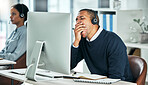Young call center agent yawning while sitting at his desk working on a computer in the office. Exhausted, tired and overworked man in financial customer service speaking to a client with his headset.