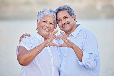 Buy stock photo A happy senior couple with a heart sign with their fingers and enjoying fresh nature air on vacation at beach while bonding. Portrait of retired couple hugging at beach with smile and love together